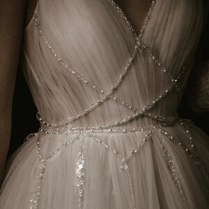 May include: A white tulle dress with a beaded bodice and a flowing skirt. The beads are arranged in a criss-cross pattern and create a delicate and elegant look.