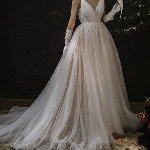 May include: A white tulle wedding dress with a V-neckline and a beaded bodice. The dress has a long train and is worn by a model holding a white bird.