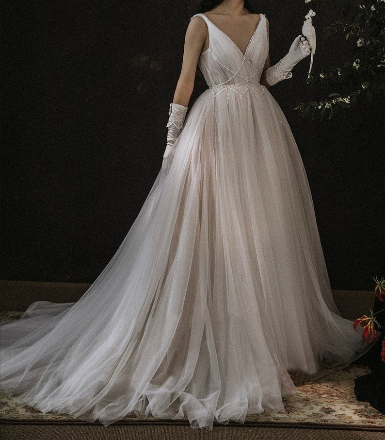 May include: A white tulle wedding dress with a V-neckline and a beaded bodice. The dress has a long train and is worn by a model holding a white bird.