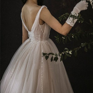 May include: A white tulle wedding dress with a low back and beaded embellishments. The dress has a full skirt and is worn by a person with white gloves.