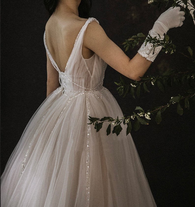 May include: A white tulle wedding dress with a low back and beaded embellishments. The dress has a full skirt and is worn by a person with white gloves.