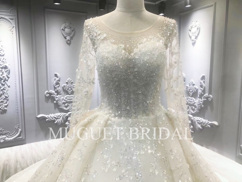 May include: A white wedding dress with long sleeves and a beaded bodice. The dress has a full skirt and is decorated with sequins and lace.