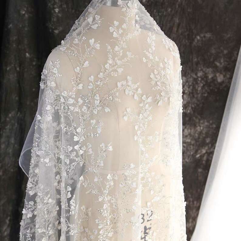 May include: White beaded lace fabric with a floral pattern. The fabric is sheer and delicate, with a soft, flowing texture.
