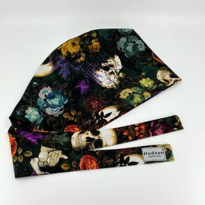 May include: A dark green scrub cap with a floral and skull pattern. The cap features roses and other flowers in shades of yellow, orange, and blue, with white skulls scattered throughout. The cap has a matching tie with the text "Hudson SCRUB CAPS".