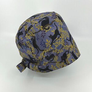 May include: A scrub cap featuring a repeating pattern of black cats perched on branches with yellow leaves against a muted purple background. The cap has a tie in the back for an adjustable fit.