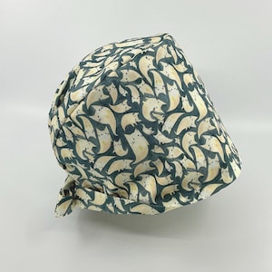 May include: A patterned surgical scrub cap featuring a repeating design of cream-colored cat silhouettes on a teal background. The cap is tied at the back and is made of a soft, flexible fabric. The cap is designed to be worn in a medical or veterinary setting.