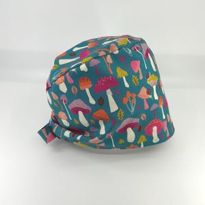 May include: A teal fabric cap featuring a colorful pattern of mushrooms in shades of pink, orange, yellow, and white. The cap has a tie closure at the back and is made of a soft, patterned fabric. The design includes various mushroom illustrations.
