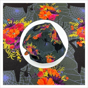 May include: A dark green surgical scrub cap featuring a floral and Halloween-themed pattern. The cap is adorned with orange, pink, and purple flowers, spiderwebs, spiders, and a witch's cauldron. The background fabric has a similar pattern.