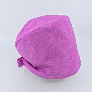 May include: A pink surgical scrub hat with a swirling, abstract pattern. The hat has a tie at the back.