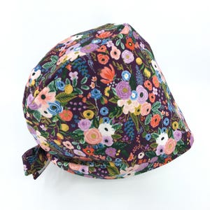 May include: A purple scrub hat with a floral print featuring pink, white, yellow, and blue flowers. The hat has a tie at the back.
