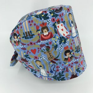 May include: A blue scrub hat with a whimsical Alice in Wonderland print featuring characters like the Queen of Hearts, the Mad Hatter, and the Cheshire Cat. The print is on a light blue background with gold accents.