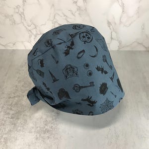 May include: A blue surgical scrub cap with a Halloween-themed pattern. The cap features black illustrations of bats, pumpkins, keys, and other spooky elements. The cap is tied at the back.