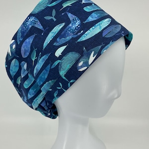May include: A scrub cap featuring a dark blue background with a repeating pattern of various whales and sea creatures in shades of light blue, teal, and white. The fabric has a textured appearance with diverse artistic whale illustrations.