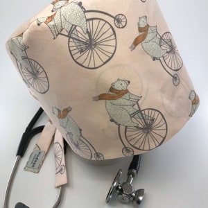 May include: A light pink medical scrub cap featuring a repeating pattern of bears riding penny-farthing bicycles. The bears wear brown scarves. A stethoscope is next to the cap.