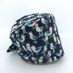 May include: A navy blue scrub cap featuring a whimsical pattern of cartoon seagulls wearing hats and boots. The seagulls are in various poses, with white, blue, red, and yellow accents. The cap is tied at the back.