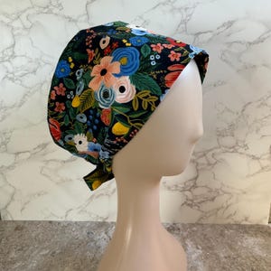 May include: A floral patterned scrub cap featuring a dark background with colorful flowers in shades of pink, blue, and yellow. The cap is tied at the back and is designed to cover the head and hair.