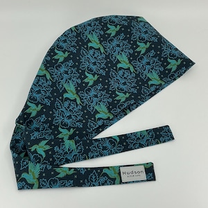 May include: A dark navy blue scrub cap featuring a repeating pattern of light blue hummingbirds and floral outlines. The fabric includes small white cross symbols throughout the design. A white rectangular label reads Hudson Scrub Caps.