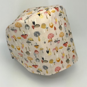 May include: A beige surgical scrub cap with a repeating pattern of colorful mushrooms in red, yellow, brown, and gray. The mushrooms are stylized and arranged on the fabric. The cap is made of a soft, likely cotton, material.
