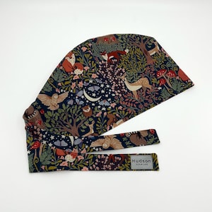 May include: A navy blue scrub cap with a woodland-themed pattern featuring foxes, deer, owls, mushrooms, and a crescent moon. The cap includes a matching tie and a tag that reads "Hudson Scrub Caps."