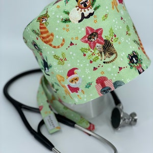 May include: A light green medical scrub cap with a festive cat print. The cap features various cats dressed in holiday attire, including Santa hats and reindeer antlers, set against a background of holly and stars. A black stethoscope rests nearby.