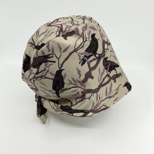 May include: A beige fabric cap featuring a repeating pattern of black crows perched on brown branches with purple leaves. The cap is tied at the back and is suitable for various uses.