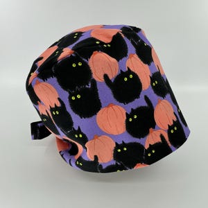 May include: A purple and black Halloween-themed scrub cap featuring a repeating pattern of black cats and orange pumpkins. The cap has a black adjustable strap and is made of fabric.