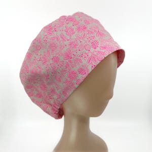 May include: A pink and white floral patterned scrub cap. The cap features a repeating design of flowers, leaves, and berries. The cap is made of a soft fabric and is designed to be worn in a medical or healthcare setting.