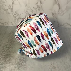 Puede incluir: Un gorro quirúrgico blanco con un patrón repetido de plumas coloridas en tonos de azul, morado, naranja y verde. El gorro está atado en la parte posterior y está hecho de una tela suave y ligera.