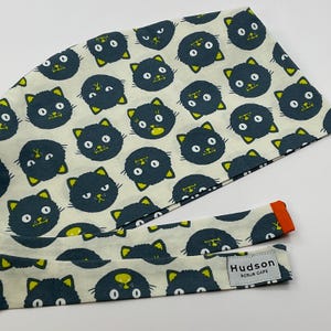 May include: A scrub cap featuring a repeating pattern of dark blue cat faces with yellow accents on a cream background. The cap has a matching tie with an orange edge. A tag reads "Hudson Scrub Caps."