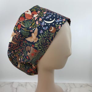May include: A dark blue scrub cap with a whimsical forest scene. The cap features illustrations of owls, butterflies, mushrooms, and a crescent moon. The design includes brown, tan, red, and pink elements.