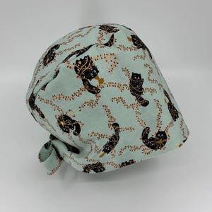 May include: A light blue surgical scrub cap with a repeating pattern of black cats and string lights. The cap is tied at the back and is made of a soft, patterned fabric. The cats are in various poses.
