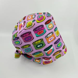 May include: A lavender surgical scrub cap featuring a repeating pattern of colorful cat food cans. The cans are in various colors, including pink, green, and orange, with fishbone designs. The cap is tied at the back.