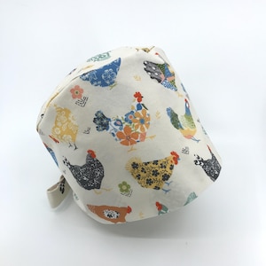 May include: A cream-colored cap featuring a repeating pattern of colorful chickens. The chickens are decorated with floral and geometric designs in shades of blue, yellow, orange, and black. The cap has a tie closure.