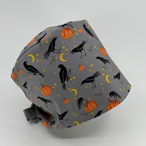 May include: A gray surgical scrub cap with a Halloween-themed pattern. The fabric features black crows, orange pumpkins, yellow crescent moons, and stars. The cap is tied at the back.