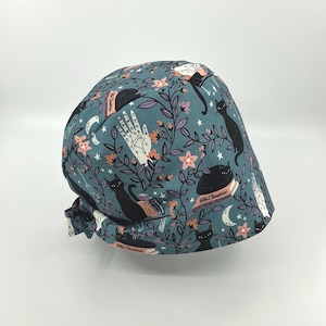 May include: A teal scrub cap with a repeating pattern of black cats, white hands, moons, flowers, and books. The cap is tied in the back and features the text "The Creatures Practical Potions".