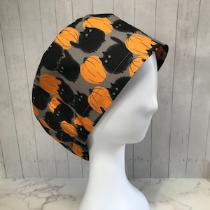 May include: A Halloween-themed surgical scrub cap featuring a repeating pattern of black cats and orange pumpkins on a gray background. The cap is displayed on a white mannequin head, showcasing its shape and design. The fabric appears to be cotton.