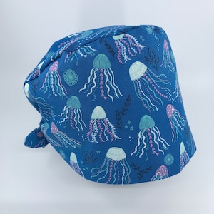 May include: A blue scrub cap featuring a repeating pattern of jellyfish in shades of teal, pink, and white. The cap is made of fabric and has a tie in the back. The design includes sea plants and stars.