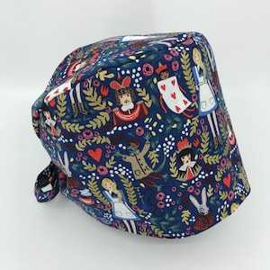 May include: A navy blue scrub cap featuring an Alice in Wonderland theme. The cap is patterned with illustrations of Alice, the Queen of Hearts, the Mad Hatter, and the White Rabbit, along with hearts and floral accents. The cap has a tie closure.