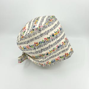 May include: A cream-colored surgical scrub cap featuring musical notes and colorful floral patterns. The cap has a tie in the back for an adjustable fit. The design includes black musical notation with vibrant flowers in shades of pink, blue, yellow, and green.