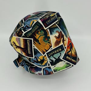May include: A bucket hat featuring a colorful collage of vintage-style travel posters depicting mythical creatures. The print includes labels such as Jackalope, Kentucky Goblin, Cave Dragon, Keelut, and Bigfoot, each paired with a national park name.
