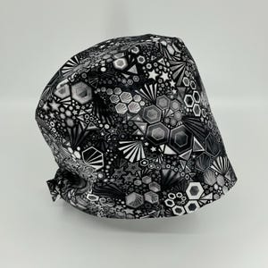 May include: A black and white patterned scrub cap. The cap features a geometric design with hexagons, stars, and other shapes. The color scheme is primarily black, white, and shades of gray. The cap is tied at the back.