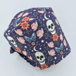 May include: A purple scrub hat with a floral pattern featuring white skulls, butterflies, and crescent moons. The flowers are pink, orange, and red.