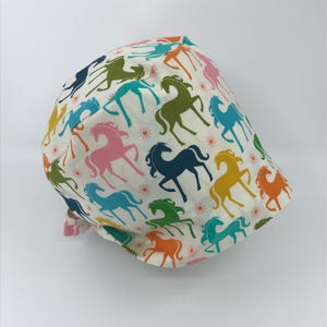 May include: A white fabric cap featuring a repeating pattern of colorful horse silhouettes in shades of pink, blue, green, yellow, and orange. Small orange flowers are interspersed throughout the design. The cap has a curved brim and a tie closure.