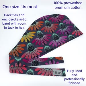 May include: A gray scrub cap with a floral print of pink, purple, orange, and yellow flowers. The cap has a tie at the back and is fully lined. The text "Hudson Scrub Caps" is printed on a white tag attached to the cap.