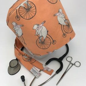 May include: A peach-colored scrub cap with a repeating pattern of bears riding penny-farthing bicycles. A matching stethoscope band and medical instruments are also present.