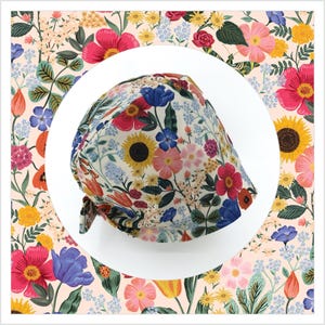 May include: A floral patterned scrub cap featuring a variety of colorful flowers, including sunflowers, tulips, and roses, on a cream background. The cap has a tie in the back and is surrounded by a matching floral pattern.