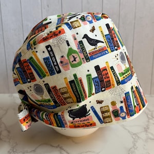 May include: A white surgical scrub cap with a colorful print of stacked books, potion bottles, spiders, and a black crow. The cap features a tie closure and is designed for medical professionals or those in the healthcare field.