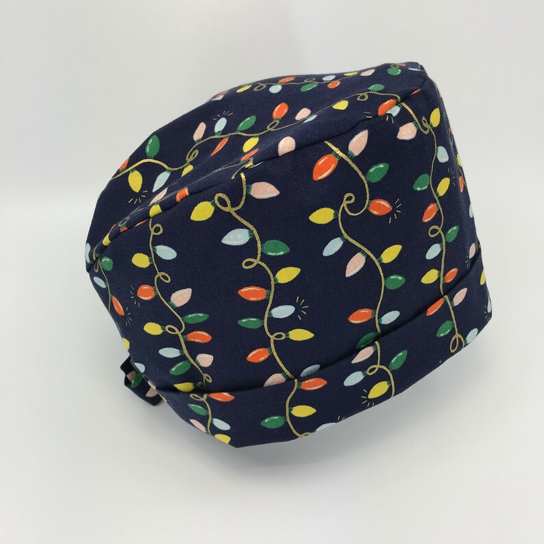 May include: A navy blue scrub hat with a pattern of colorful Christmas lights. The hat has a tie on the back.