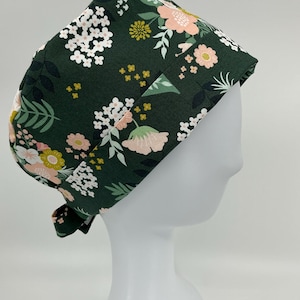 May include: A scrub cap featuring a dark forest green background with a floral pattern of white, light pink, and mustard yellow flowers and green leaves. The cap has a folded brim and a tie-back closure, providing a secure fit for the wearer.