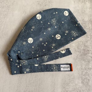 May include: A dark blue scrub cap with a celestial pattern of white stars, moons, and bursts. The cap has a long tie and a small tag that reads "Hudson Scrub Caps" with an orange border.